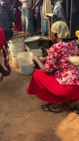 This is a street scene at a market. The ground is dirt with some puddles and debris. In the foreground, a person wearing a red shirt and black shorts is bent over, holding a bundle of dark-colored hair. To the right, another person is seated, wearing a red skirt. In the background, several people are walking and standing. One person in a black and white shirt and leggings is carrying a white bag and a black bag. Another person is wearing a patterned dress. There are market stalls with various items, including buckets, produce, and textiles. A young child is standing near a cart laden with goods. An older man is gesturing with his hands.