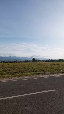 The video shows a roadside view of a field against a mountain range. The sky is light blue with some clouds. The field is mostly brown and green grass. A two-lane asphalt road with a white line divider runs along the bottom of the frame. Some buildings are visible in the distance between the field and the mountains. Unidentifiable chatter can be heard in the background. The camera remains static throughout the video.