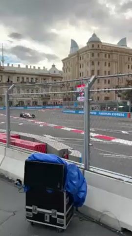 The video shows a race car speeding along a track surrounded by barriers and sponsor advertisements. The architecture of a large, ornate building with a domed roof serves as the backdrop. A crowd of spectators lines the street behind a fence, and a section of colorful stadium seating is visible. The sky is overcast.