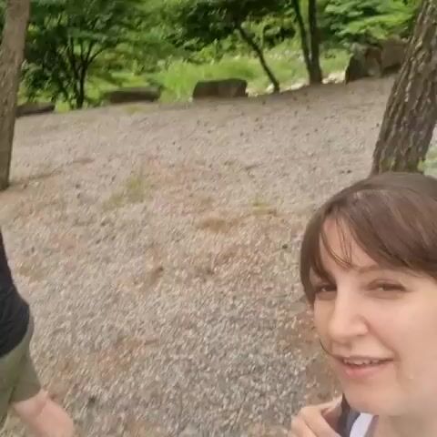 A low-resolution video captures a family in an outdoor environment, likely a park or wooded area. The ground is covered with gravel and fallen leaves, with trees and foliage in the background. In the foreground, a woman with brown hair and bangs is facing the camera and smiling. Behind her, a man in a black polo shirt and olive green shorts is carrying a young child, who is wearing a black and white shirt, on his left hip. The child is facing away from the camera and appears to be nuzzling the man's neck. The video is grainy and somewhat blurry, suggesting it was taken with a low-quality camera or in low light conditions.