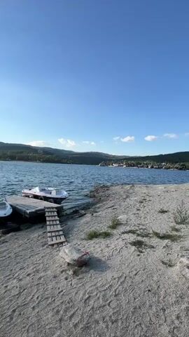 The video shows a sandy shore with a lake in the background. Two boats are docked on a wooden pier. A small wooden ramp leads from the shore to the pier. The sky is blue with some clouds. The sun is shining and reflecting on the water. In the distance, there are hills covered with trees and some buildings. There are no people visible in the video.