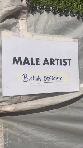 The video shows what appears to be a costume or wardrobe department on a film set or production.  The initial shot shows a sign indicating the role of "Male Artist" with a handwritten addition reading "British Officer." The camera then pans to reveal a white tent-like structure, and then enters the tent, where several people are present. Some are looking at clothing, others are sitting down, and one person is using an iron.  Various pieces of clothing are on display, including a long brown garment hanging on a hanger. The setting appears to be outdoors, with trees visible in the background. There are also some yellow plastic crates and metal surfaces present.