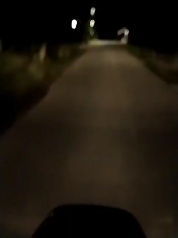 The video depicts a nighttime scene, likely captured from a moving vehicle on a paved road. Streetlights illuminate the road ahead, casting a bright, diffused glow. The road appears relatively straight and empty, with sparse vegetation and buildings visible on either side. The overall atmosphere is dark and quiet.