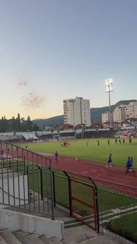 A wide shot of a soccer stadium at dusk, with a clear sky and scattered clouds. A soccer game is in progress on the green field, with players in yellow and blue uniforms visible. There are spectators in the stands. In the background, there are tall buildings and hills. To the left, there is a small display of fireworks. The camera is positioned in the stands, looking down at the field.