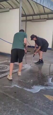 Two individuals are shown cleaning a rug with pressure washers. The person on the left, wearing a green t-shirt and black shorts, operates a pressure washer directed at the rug. The person on the right, wearing a black t-shirt and black shorts, is also using a pressure washer on the rug. The rug itself is patterned with concentric circles and a geometric border. The ground is wet and foamy, indicating the cleaning process is underway. They are in an open, covered area, possibly an outdoor car wash or a similar facility, with a wall and a metal roof structure visible. The lighting suggests daylight.