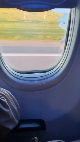 The video shows a view from an airplane window during takeoff. The camera is positioned inside the cabin, looking out at the runway and the surrounding landscape. The airplane's wing is visible in the upper right corner of the frame, and the runway lights and markings are clearly discernible. The ground outside is a blur of green fields and a strip of pavement, indicating rapid movement. The inside of the airplane cabin is visible in the foreground, with a seatback and a pull-down shade obscuring parts of the view. The lighting suggests that it is either early morning or late afternoon, with the sun casting a warm glow on the scene.