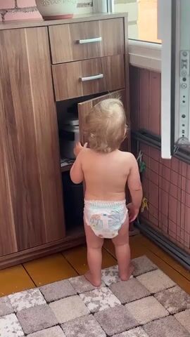 A toddler stands facing a wooden cabinet with one door open. The toddler is wearing a diaper and has blonde curly hair. The cabinet has three drawers at the top and a single door that is currently open, revealing the inside. The toddler is reaching towards the open cabinet door. The floor is covered with a checkered rug. The background shows a pink wall, a window, and a piece of furniture with a white front.