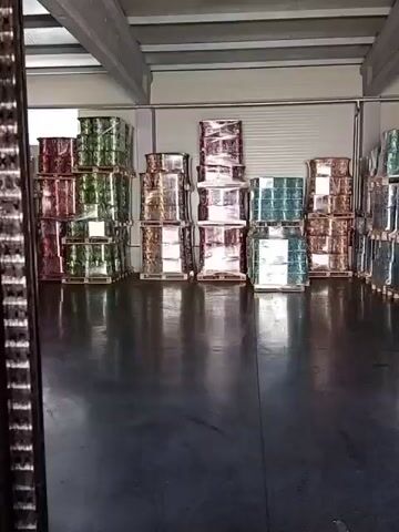 The video shows the interior of a warehouse. The floor is a dark, reflective material, likely concrete. The walls are light-colored metal. The ceiling is also made of metal beams and panels. Pallets of goods are stacked throughout the warehouse. Some pallets are wrapped in clear plastic and contain items that appear to be drums or large containers. These are stacked in rows. Other pallets contain cardboard boxes, some stacked, others in smaller piles. The lighting is from overhead fixtures. The video appears to be filmed from a moving perspective, likely from a forklift or similar vehicle, as suggested by the close-up of what appears to be part of a forklift's structure at the beginning and end of the clip. The overall impression is one of organized storage of goods in a commercial or industrial setting.