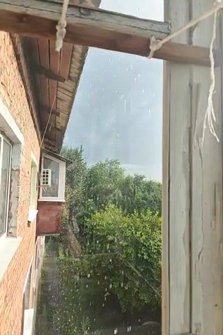 The video shows a view through a window, with raindrops visible on the glass. The scene outside is a suburban landscape with houses and trees. The sky is cloudy with the sun peeking through, suggesting it might be during or just after a rain shower. The overall mood appears calm and somewhat melancholic due to the rainy weather. The architectural style of the houses suggests a European setting. There is no visible text in the video.