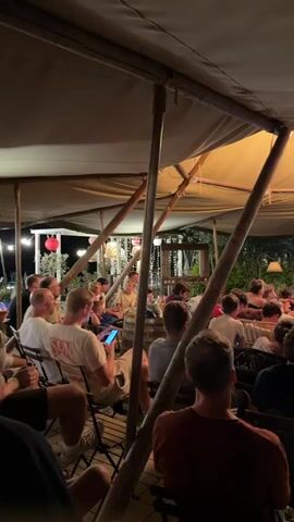 The video shows a group of people gathered under a large tent or canopy structure. They are seated in chairs and on cushions, facing a television screen that is displaying a soccer match. The lighting is dim, with warm light coming from overhead fixtures, including a chandelier. The environment appears to be outdoors, possibly a patio or garden area, with greenery visible in the background. Some individuals are holding drinks or looking at their phones. The overall atmosphere suggests a social gathering for watching a sporting event.