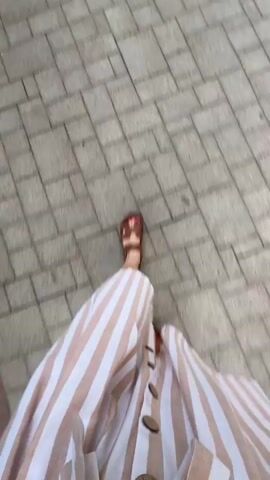 The video shows a first-person perspective of someone walking on a paved sidewalk. The pavement is made of light gray square tiles arranged in a repeating pattern. A person is wearing a striped dress with vertical lines of white and light brown, and brown sandals. The person's legs and feet are visible as they walk. The camera angle is looking down at the ground. The lighting suggests an outdoor environment.