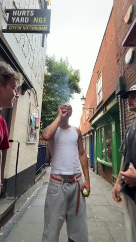 In a narrow alleyway marked "Hurts Yard NG1, City of Nottingham," three young men are standing together. The man in the center, wearing a white tank top and gray shorts with an orange belt, is focused on something in his hands. He appears to be manipulating or examining a small, dark object. He is flanked by two other men. The man on the left is wearing a red t-shirt and gray shorts and is looking at the man in the center. The man on the right is wearing a black patterned t-shirt, white shorts, and a white baseball cap, and is partially obscured. The alleyway is flanked by brick and white-painted buildings, with signs visible for a barbershop and a restaurant. A lamppost is visible in the background.