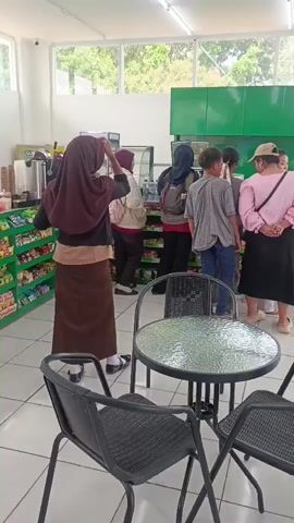 This video shows a group of people inside a brightly lit convenience store. In the foreground, a round glass-topped table with four black chairs surrounds it. In the background, shelves are stocked with various products. A woman in a brown headscarf and long skirt stands on the left, facing away from the camera. Next to her, another person in a blue headscarf is visible. A group of three people, two males and one female, stand in the center, looking towards the counter. On the right, a person in a pink shirt and black skirt stands with their back to the camera, and a woman in a red shirt is interacting with someone behind the counter. The overall environment appears clean and organized.