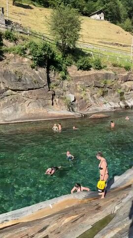 This media file captures a sunny day in Verzasca, Switzerland, at a river known for its emerald green waters. The scene is a natural swimming area where several people are enjoying the refreshing water. In the foreground, a woman in a black bikini stands on a large, smooth rock, holding a bright yellow waterproof bag. Another person is partially submerged in the water near her, and further back, more individuals are swimming and relaxing in the river.

The river is flanked by dramatic, layered rock formations and steep, tree-covered hillsides, a characteristic of the Verzasca Valley. A rustic stone building is visible on a grassy slope in the upper background, along with a fence line and a dirt path. The water is remarkably clear, allowing a view of the rocky riverbed beneath. The bright sunshine casts shadows, indicating it is likely midday or early afternoon. The overall atmosphere appears relaxed and joyful, with people taking advantage of the warm weather and beautiful natural environment. There is no visible text within the media file.