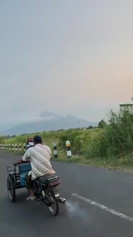 A person wearing a blue baseball cap and a striped shirt is riding a three-wheeled motorcycle with a cargo box attached to the back. The motorcycle is on an asphalt road with a white dashed line in the center. There is smoke coming from the exhaust pipe of the motorcycle. In the background, there are green fields with tall grass and some trees. In the distance, there are mountains under a hazy sky. Another motorcycle with two passengers is visible further down the road.