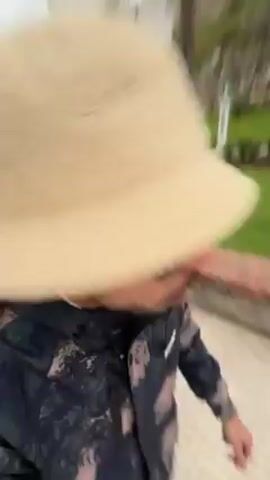 The video shows a person wearing a cream-colored bucket hat, sunglasses, and a patterned jacket walking on a paved pathway. Behind the person is a green grassy park area with palm trees, statues, and a brick border along the pathway. On a hill in the background, there is a stone building or castle. The sky is overcast. The camera appears to be handheld and is recording from the perspective of someone walking alongside the person.