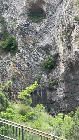A person is suspended by a rope system, swinging horizontally across a massive, grey, striated rock cliff face. The person wears light-colored pants and a dark top with a visible yellow or light-colored harness. Below the swinging person and in the foreground, green trees and foliage are visible. A metal bridge with a railing is partially seen at the bottom of the frame. The person swings from the left side of the frame towards the right, moving past the distinct geological layers of the cliff.