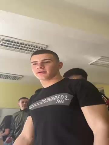 A young man wearing a black t-shirt with white lettering "DSQUARED2 1964" is speaking in front of a group of people in a room. He has short hair and his head is tilted slightly up and to the left. In the background, there are other people, including a young man in a green jacket and a person with a hood. The room has a yellow wall, a fluorescent light fixture, and a projector screen.