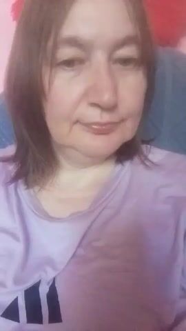 A woman with shoulder-length brown hair is seen in a close-up shot. She is wearing a light purple t-shirt with a logo on it. The background is blurred but appears to be a room with pink and blue elements. The woman looks at the camera and smiles, then brings her hand to her mouth as if to kiss.
