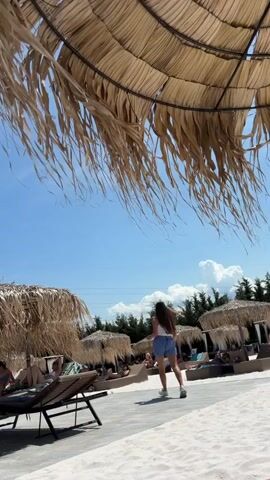 A young woman with long brown hair, wearing a white tank top and blue shorts, walks across a sandy area. She is holding a tray with what appears to be a drink or food on it. She is walking away from the camera. In the background, there are several thatched-roof umbrellas providing shade over lounge chairs where other people are sitting or lying down. The sky is blue with some white clouds. There are trees visible in the distance. The scene appears to be a beach or poolside resort.