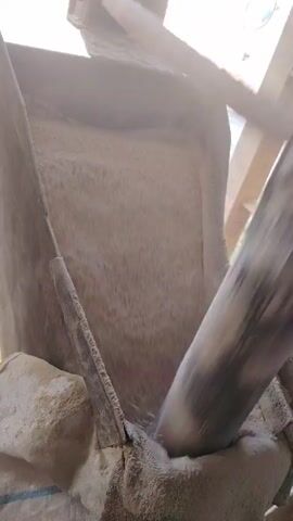 The video shows a close-up of a mixing process inside a large container. A blunt, angled arm repeatedly rotates within the container, stirring and mixing its contents, which appear to be a fine, light-colored, grainy substance. The interior of the container is rough and textured. The environment seems to be outdoors with natural light.