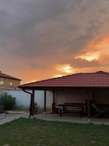 The video pans across an outdoor scene at dusk. In the foreground, a grassy lawn leads to a covered patio area with a red tiled roof. Under the patio, there is a wooden table and benches, a stroller, and a hanging swing chair. To the right of the patio, a house constructed of concrete blocks with a red tiled roof is visible, featuring an open doorway. The sky above is filled with dramatic clouds, illuminated by the warm oranges and pinks of a sunset. The overall impression is a peaceful, residential setting during a beautiful sunset.
