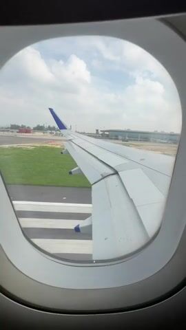 The video shows a view from an airplane window during takeoff. The wing of the plane is visible, along with a runway and an airport terminal in the background. The sky is partly cloudy. The plane appears to be accelerating down the runway.