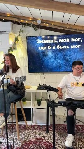 The video shows a band performing for an audience in a room. The band consists of a female singer and guitarist, a second female singer, two guitar players, and a percussionist. There is a large TV screen behind the band, and a rug on the floor. The audience is made up of people of all ages, including children. Some of the audience members are seated, while others are standing. The room appears to be well-lit and decorated with plants.