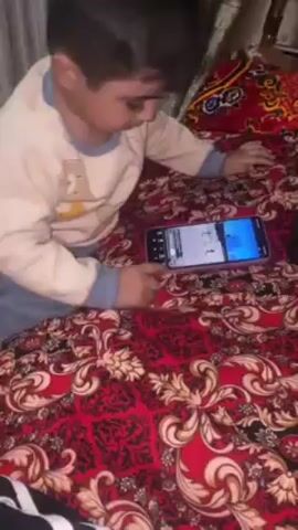 A toddler is lying on their stomach on a bed, looking at a cell phone. The bedspread is red with a busy pattern of dark red, gold and white. The toddler is wearing a light-colored, long-sleeved shirt and blue pants. They appear to be watching a video on the phone, and they point to the screen while looking up and yelling. A black and white striped object is visible on the lower left corner of the frame. Curtains are visible in the background behind the head of the bed.