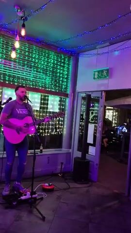 A man is playing an acoustic guitar and singing into a microphone in what appears to be a bar or restaurant. He is wearing a grey T-shirt with the word "Vans" on it, blue jeans, and sneakers.  The lighting in the room alternates between red, green, blue, and purple, and there are string lights along the ceiling. Behind him is a large window with blinds and more string lights. To his right, there is an open doorway leading to another room with tables and chairs. A speaker and some sound equipment are on the floor near him. The man is looking directly into the microphone as he plays and sings.