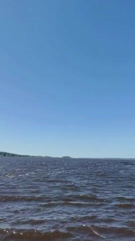 The media file depicts a wide, choppy body of water under a clear, bright blue sky. The water is a dark, reddish-brown color, suggesting it might be a lake or a slow-moving river, with small whitecaps indicating a light breeze. In the distance, a hazy shoreline is visible, dotted with indistinct structures that could be buildings or trees. A small, dark shape, possibly a distant island or a boat, can be seen on the horizon to the right. The overall impression is of a calm, sunny day, likely in the late morning or early afternoon, given the bright light and clear sky. There is no visible text in the media file. The scene conveys a sense of vastness and natural beauty, with the expansive sky and water dominating the frame.