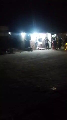 The video shows a brightly lit shop at night. People are standing and milling around outside the shop. A motorcycle is visible among them. The ground is dark and rough. The camera moves slightly during the recording. The overall impression is of a gathering at a local store at night.