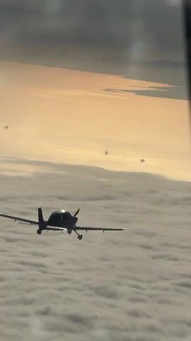 A small propeller airplane flies above a layer of clouds. The sun is setting and casts a golden glow on the horizon. Several other small airplanes are visible in the distance, flying in formation. The image is framed by a dark, blurry object in the foreground, possibly the wing or strut of another aircraft.