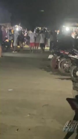 At night, a crowd gathers along a road lined with parked motorcycles. Some individuals stand near a light source, while others walk along the road. The road surface appears uneven. The license plate on a motorcycle in the foreground reads L5701. The background is dark and indistinct.