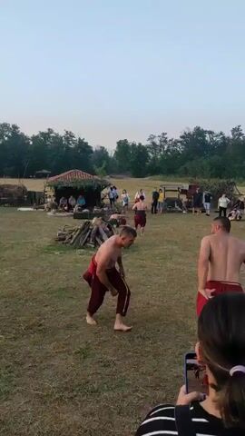 The video depicts a wrestling match in an outdoor grassy area. Two shirtless men wearing red baggy pants engage in a wrestling match. One man attempts a leg sweep but the other man dodges. They then grapple, moving across the field. In the background, there is a small thatched-roof building, and several people are seated or standing, watching the match. There are also piles of logs and various items scattered around the field, suggesting a rural or historical setting. The sky is overcast and bright. The video is filmed from the perspective of someone holding a phone, with a partial view of the phone and the person's hand visible in the lower right corner.
