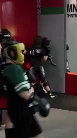 The video shows a group of children practicing martial arts. Three children are wearing helmets and boxing gloves, while others are standing in the background. The children are sparring near a padded red wall in what appears to be a gym or training facility. They are throwing punches and kicks, attempting to strike each other. The room is lit and appears to be indoors.