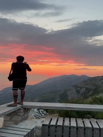 The video shows a person with dark hair and dark clothing, wearing shorts and sandals, standing on a stone ledge and taking a photo of a sunset with their phone. The sun is a bright red orb setting behind a mountain range, with clouds illuminated by its rays in shades of pink and orange. The landscape is mountainous with green vegetation. A wooden bench is visible in the foreground.