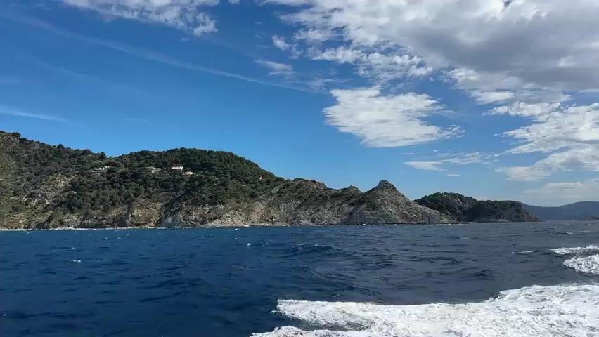This video captures a view from a moving boat on a bright, sunny day with a blue sky dotted with white clouds. The boat is creating a wake of white water on the right side of the frame. In the distance, a rugged, tree-covered island or coastline is visible. The land rises steeply from the sea, with rocky cliffs and dense green vegetation. A prominent white structure, possibly a lighthouse or a building, is situated on the highest peak of the island. Scattered houses are also visible amongst the trees on the slopes. The sea is a deep blue and appears somewhat choppy.