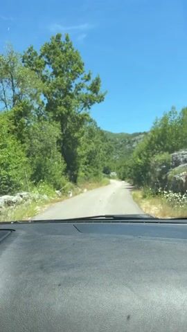 A video taken from inside a vehicle shows a narrow, paved road winding through a wooded area. The road is bordered by lush green trees and vegetation. The sky is a clear blue with a few wispy clouds. The vehicle appears to be moving forward, indicated by the slight blur of the trees and the perspective of the road. The dashboard of the vehicle is visible in the foreground.