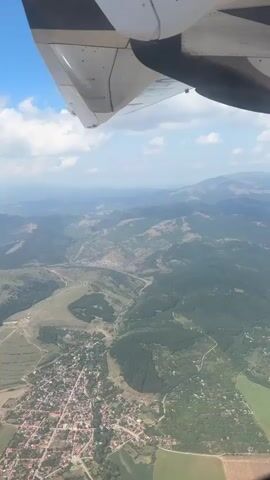 The video provides an aerial view from an airplane window, looking down at a rural landscape. The edge of the airplane wing is visible in the foreground, with white and black stripes. Below, rolling hills covered in green trees and vegetation dominate the scene. A small town or village with red-tiled roofs is nestled in a valley, with roads winding through it and connecting to the surrounding countryside. Patches of farmland, some green and some appearing tilled, are interspersed with the trees. In the distance, more hills and mountains fade into a hazy blue sky dotted with white clouds. The overall impression is of a serene, natural environment seen from high above.