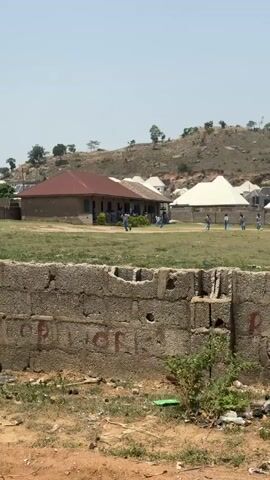 The video shows an outdoor scene in Suleija, Nigeria, during the daytime under a clear, bright sky. In the foreground, there is a low, unfinished concrete block wall with faded red graffiti, where parts of letters like "S," "P," "O," and possibly "R" or "J" are visible. Beyond the wall is a large, dry grassy field.

Several individuals, possibly children or young adults, wearing light blue tops and white bottoms (likely school uniforms), are seen running or walking across the field. Some are near a single-story building with a red roof on the left, while others are spread further across the field towards the right. In the middle-right, a small herd of white sheep or goats is grazing or moving.

In the background, there are several simple buildings with white or light-colored pyramid-shaped roofs, typical of residential structures. Behind these buildings, a prominent, rocky hill covered with sparse vegetation and trees rises into the distance. The overall impression is a vibrant community or school area situated near natural terrain.
