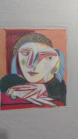 A video displays a painting of a woman's face in a cubist style. The woman has two faces, one looking forward and one in profile. She is wearing a green hat and a dark green top. Her hands are resting on a red surface. The painting is framed by a rough white border. The video pans slightly across the painting.