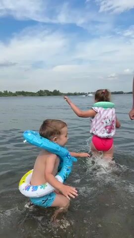 A young boy wearing a blue whale-shaped inflatable ring around his neck and a blue swim trunk is standing in shallow water on a lake, looking to his right with a smile. A young girl in a pink and white life jacket with a cat design and pink bottoms is walking further into the water, looking towards a jet ski in the distance. The water is calm with some ripples, and the sky is partly cloudy with blue patches. Trees line the distant shore.