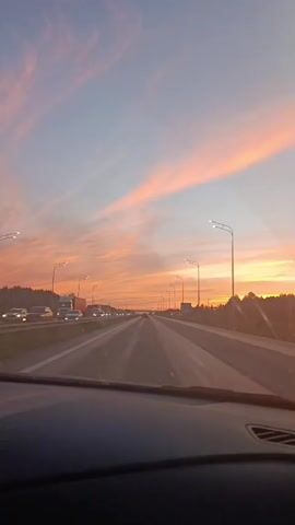 The video shows a view from inside a car driving on a highway at sunset. The sky is filled with vibrant orange and pink clouds, and the sun is setting on the horizon, casting a warm glow. The highway has multiple lanes with cars and trucks driving in both directions. Streetlights line the side of the road. The dashboard of the car is visible in the foreground.