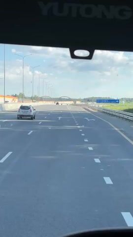 The video shows a view from inside a vehicle, looking forward through the windshield. The vehicle is on a paved highway with multiple lanes. Lane markings, including white arrows and dashed lines, are visible on the road. To the left, a white SUV is driving in the same direction. Further ahead, there are other vehicles on the highway and an arched overpass. On the right side of the highway, there are traffic signs, lampposts, and grassy areas with trees in the distance. The sky is cloudy. The word YUTONG is visible on the top interior of the windshield.