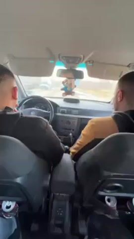 The camera is positioned in the back seat of a car, looking forward between the two front seats. Two men are in the front seats, the driver and the passenger. They both have short hair and are facing forward. The driver is wearing a dark shirt, and the passenger is wearing a yellow and black jacket. The car is driving down a road with green trees on either side. There is a brown stuffed animal hanging from the rearview mirror. There are empty cans visible in the back of both front seats. The dashboard of the car is visible, including the steering wheel, radio, and climate controls. The sun visors are pulled down.