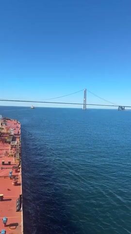 The video shows a large, red-decked ship with numerous pipes and equipment on its surface sailing on a vast body of blue water under a clear blue sky. The ship is seen from a side-on perspective, with its bow facing away from the camera. In the distance, a large suspension bridge stretches across the water, with its towers clearly visible. Another smaller vessel is also visible on the water further away from the bridge. The camera appears to be positioned on another vessel or elevated platform, looking down and across at the ship and the bridge. The overall scene depicts a maritime journey with a significant bridge in the background.