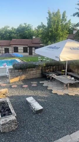 A man is walking into frame from the right carrying a package of drinks. He is wearing a white t-shirt. He is in an outdoor area with a swimming pool, picnic tables, and a fire pit. The area is surrounded by trees.