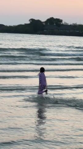 A low-angle shot shows a beach scene at dusk. In the foreground, a child wearing a purple, long-sleeved swimsuit and a black head covering is running into the ocean, kicking up splashes. Their reflection is visible in the water. The ocean has gentle waves, and several people are swimming in the distance. Behind them, there is a small island with trees and what appears to be a building. The sky is a light pink and grey hue, suggesting it is either dawn or dusk.