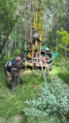 Two men are working near a drilling rig in a wooded area. One man is crouched down and appears to be operating a piece of equipment near the base of the rig, while the other man stands nearby observing. The drilling rig is yellow and green and appears to be mounted on a vehicle. The environment is lush with green trees and grass.